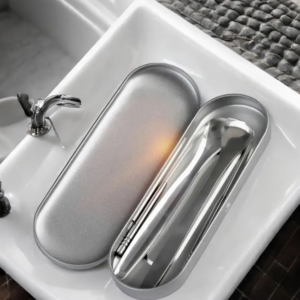 Kit limpiador de lengua de acero quirúrgico plateado con estuche metálico ovalado, mostrando dos raspadores metálicos y un palillo en su interior sobre un lavabo blanco.